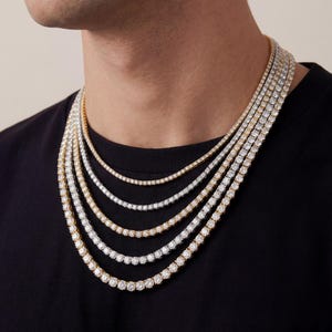 May include: A layered set of five tennis necklaces featuring round-cut clear stones set in alternating gold and silver-toned metal chains. The necklaces are arranged in a cascading style, each displaying a continuous row of sparkling stones.