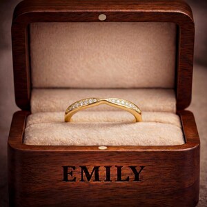 May include: A gold ring with a curved band featuring a row of small clear stones, presented inside a dark brown wooden box with a beige velvet interior. The front of the wooden box is engraved with the name EMILY in bold, capital letters.