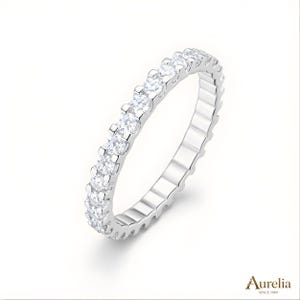 May include: A silver eternity ring featuring a continuous line of clear, round gemstones. The ring is set against a white backdrop. The brand name "Aurelia" is visible in the bottom right corner.