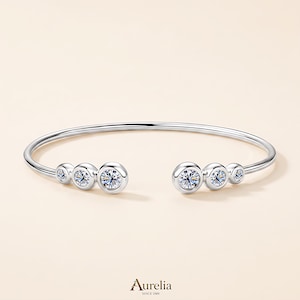Può includere: Un bracciale aperto color argento con sei pietre rotonde trasparenti incastonate a lunetta di dimensioni variabili, con tre pietre disposte su ciascuna estremità. Il nome del marchio Aurelia Since 1989 è stampato in piccolo testo nero in basso.