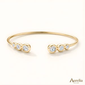 Pode incluir: Pulseira aberta dourada Aurelia com cinco pedras preciosas redondas e transparentes. A pulseira tem um design simples e elegante com as pedras engastadas em bisel. A marca "Aurelia" está impressa.