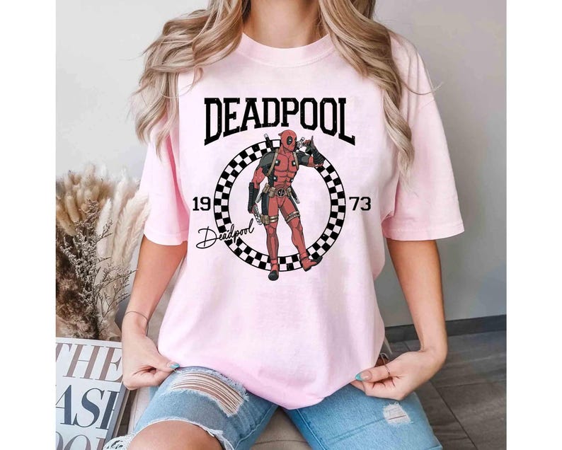 Puede incluir: Camiseta rosa claro con la palabra "DEADPOOL" en letras negras en bloque sobre un gr&aacute;fico circular. El gr&aacute;fico incluye una ilustraci&oacute;n del personaje Deadpool, los n&uacute;meros "19" y "73" y la palabra "Deadpool" en cursiva.