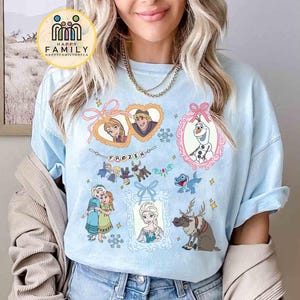 Op de afbeelding: Een lichtblauw t-shirt met een grafische print van personages uit de film Frozen, waaronder Anna, Elsa, Olaf, Kristoff en Sven. Het ontwerp bevat hartvormige kaders, een banner met de tekst FROZEN en accenten van sneeuwvlokken.