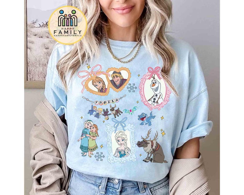 Op de afbeelding: Een lichtblauw t-shirt met een grafische print van personages uit de film Frozen, waaronder Anna, Elsa, Olaf, Kristoff en Sven. Het ontwerp bevat hartvormige kaders, een banner met de tekst FROZEN en accenten van sneeuwvlokken.
