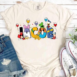 May include: A cream-coloured t-shirt featuring the name "JACOB" in vibrant, cartoon lettering. Disney characters, including Mickey and Minnie Mouse, Donald Duck, and Goofy, decorate each letter. The design is completed with balloons and stars.