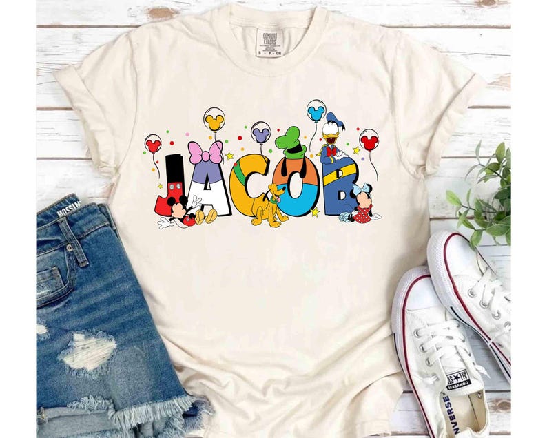 May include: A cream-coloured t-shirt featuring the name "JACOB" in vibrant, cartoon lettering. Disney characters, including Mickey and Minnie Mouse, Donald Duck, and Goofy, decorate each letter. The design is completed with balloons and stars.
