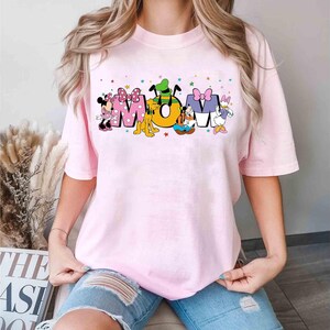 May include: A pale pink t-shirt featuring the word "MOM" in a colourful design with Disney characters. Minnie Mouse, Pluto, and Daisy Duck are included in the design. The shirt has short sleeves and a round neck.