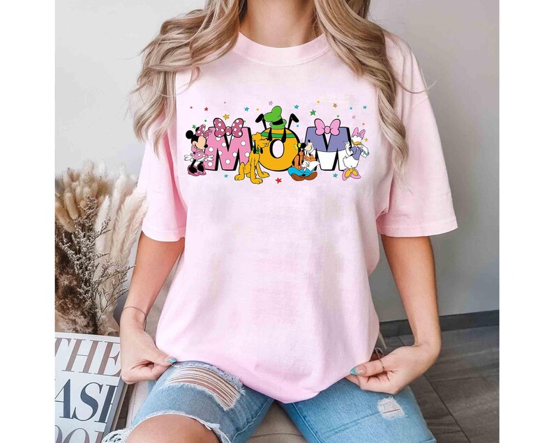 May include: A pale pink t-shirt featuring the word "MOM" in a colourful design with Disney characters. Minnie Mouse, Pluto, and Daisy Duck are included in the design. The shirt has short sleeves and a round neck.