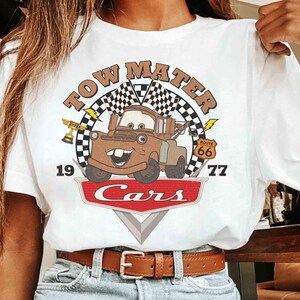 May include: White t-shirt featuring a graphic of Tow Mater from the Cars movie. The design includes the text "TOW MATER" above a cartoon image of Mater, with "1977" and "Cars" below. A Route 66 sign is also included.