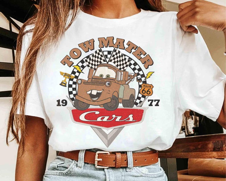 May include: White t-shirt featuring a graphic of Tow Mater from the Cars movie. The design includes the text "TOW MATER" above a cartoon image of Mater, with "1977" and "Cars" below. A Route 66 sign is also included.
