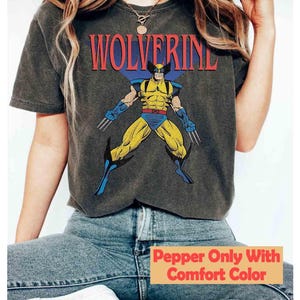 May include: A charcoal gray short-sleeve t-shirt featuring a graphic print of the comic book character Wolverine in a yellow and blue suit with metal claws. The word WOLVERINE is printed in red capital letters above the character illustration.