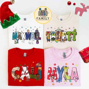 May include: Four t-shirts with personalized names and cartoon characters. The names "MAXWELL", "TRUETT", "CAILI", and "MYLA" are displayed in colourful, playful fonts. The shirts are white, beige, red, and pink. A Christmas elf hat and ornaments are in the background.