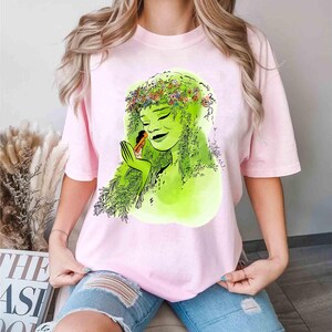 May include: A light pink t-shirt featuring a green illustration of a woman with green hair and a floral crown. The woman is holding a small figure. The shirt is a soft, comfortable material.