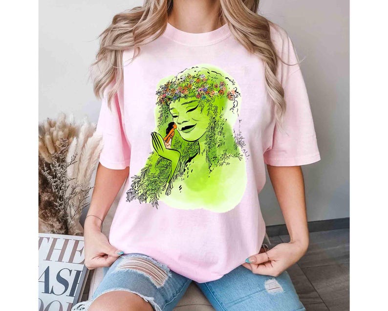 May include: A light pink t-shirt featuring a green illustration of a woman with green hair and a floral crown. The woman is holding a small figure. The shirt is a soft, comfortable material.