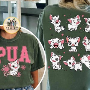 May include: Green t-shirt with the word "PUA" in pink block letters and a cartoon pig illustration. The back of the shirt features multiple cartoon pig illustrations and pink stars. The shirt is made of a soft material.