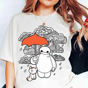 May include: A cream-colored t-shirt featuring a graphic print of a white robot holding a bright orange umbrella, accompanied by a small cat. The design includes black and white line-art clouds with intricate patterns and falling raindrops.