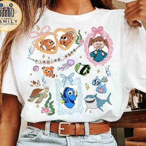 Peut inclure: Un t-shirt blanc à manches courtes avec une impression graphique colorée représentant des personnages du film Le Monde de Nemo, dont Nemo, Dory, Bruce le requin et Darla, entourés de cadres décoratifs, de bulles et d'illustrations marines.