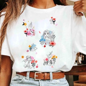 May include: White t-shirt featuring colorful cartoon illustrations of characters surrounded by flowers. The graphics include a princess, a snowman, and other characters. The shirt has a brown belt and blue jeans.