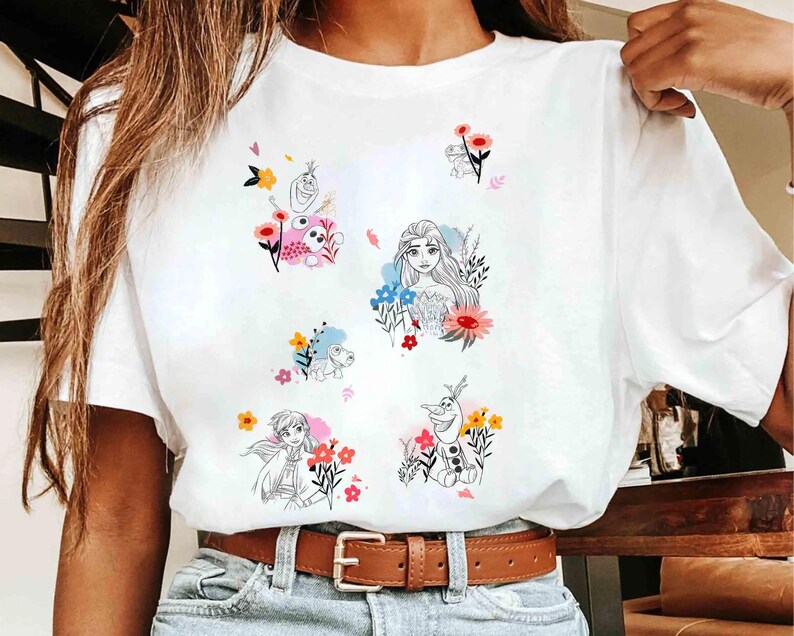 May include: White t-shirt featuring colorful cartoon illustrations of characters surrounded by flowers. The graphics include a princess, a snowman, and other characters. The shirt has a brown belt and blue jeans.
