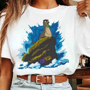 May include: White t-shirt featuring a cartoon seal holding a green bottle, perched on a rock with blue water and a purple flower. The seal has a surprised expression. The shirt is worn by a person.