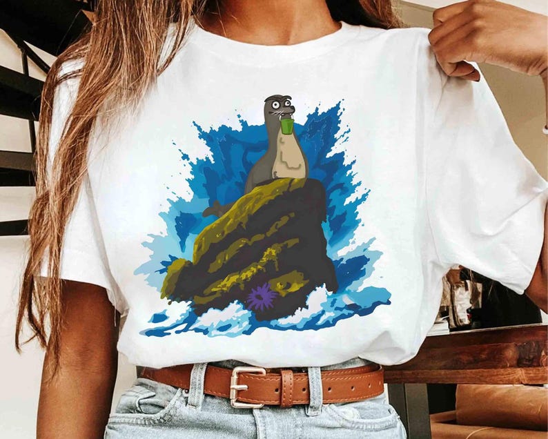 May include: White t-shirt featuring a cartoon seal holding a green bottle, perched on a rock with blue water and a purple flower. The seal has a surprised expression. The shirt is worn by a person.
