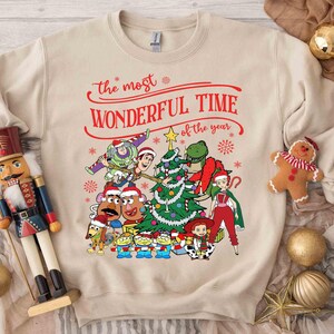 May include: Beige sweatshirt with a colorful graphic featuring Toy Story characters decorating a Christmas tree. The text "the most WONDERFUL TIME of the year" is written in red script above the graphic. A festive holiday design.
