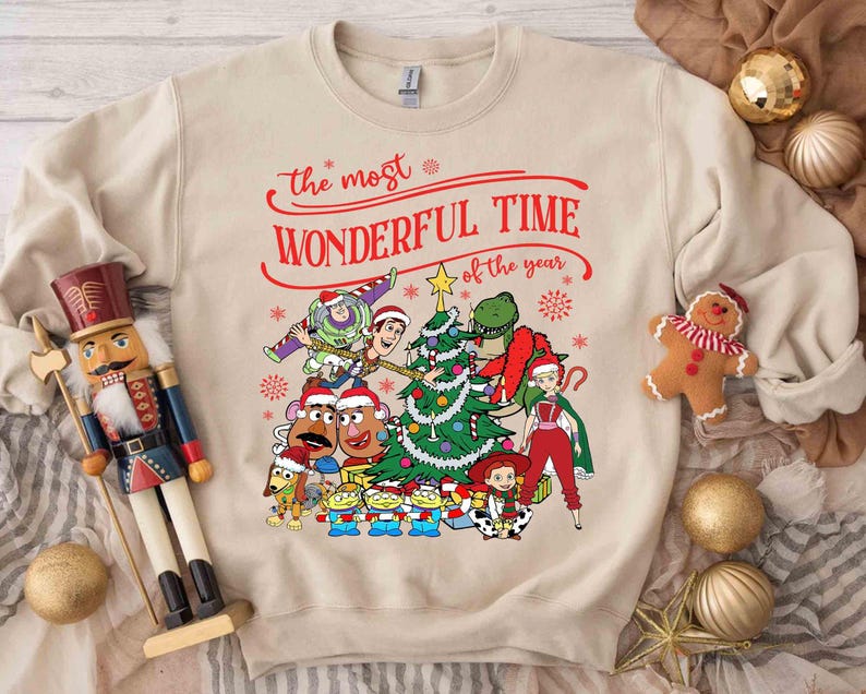 May include: Beige sweatshirt with a colorful graphic featuring Toy Story characters decorating a Christmas tree. The text "the most WONDERFUL TIME of the year" is written in red script above the graphic. A festive holiday design.