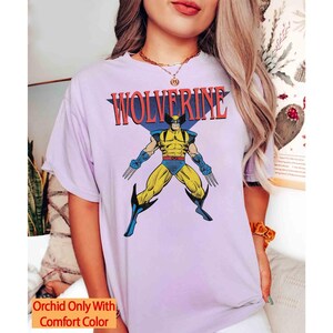 May include: A light purple short-sleeved t-shirt featuring a graphic of the comic book character Wolverine in a yellow and blue suit with extended claws. The text WOLVERINE is printed in red above the character. The shirt includes a small orange text box.