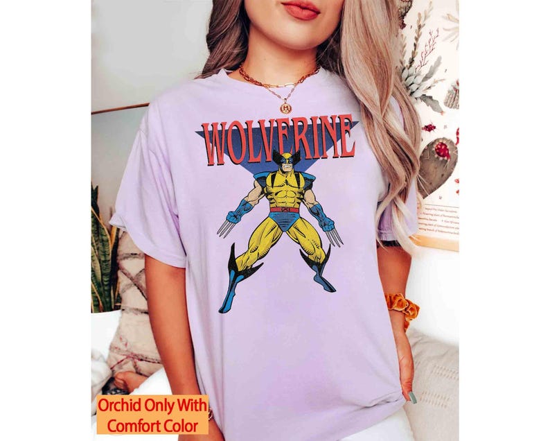 May include: A light purple short-sleeved t-shirt featuring a graphic of the comic book character Wolverine in a yellow and blue suit with extended claws. The text WOLVERINE is printed in red above the character. The shirt includes a small orange text box.