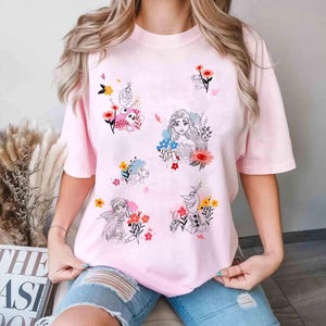 May include: A light pink t-shirt featuring illustrations of animated characters and flowers. The design includes images of a princess, a snowman, and other characters, surrounded by colorful floral accents. The shirt has short sleeves and a relaxed fit.