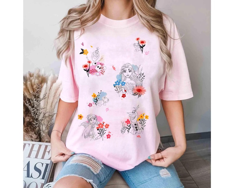 May include: A light pink t-shirt featuring illustrations of animated characters and flowers. The design includes images of a princess, a snowman, and other characters, surrounded by colorful floral accents. The shirt has short sleeves and a relaxed fit.