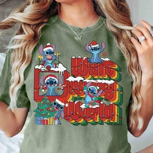 K&ouml;nnte beinhalten: Olivgr&uuml;nes T-Shirt mit einem festlichen Design mit Stitch-Charakteren mit Weihnachtsm&uuml;tzen. Die Grafik enth&auml;lt einen Weihnachtsbaum und den Text "Walt Disney World" in einer Regenbogen-Schriftart.