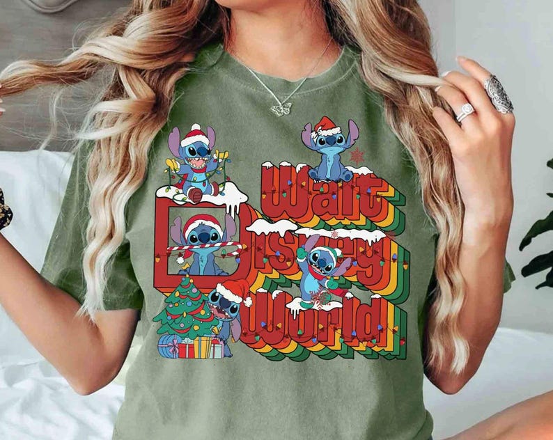 K&ouml;nnte beinhalten: Olivgr&uuml;nes T-Shirt mit einem festlichen Design mit Stitch-Charakteren mit Weihnachtsm&uuml;tzen. Die Grafik enth&auml;lt einen Weihnachtsbaum und den Text "Walt Disney World" in einer Regenbogen-Schriftart.