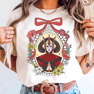Könnte beinhalten: Cremefarbenes T-Shirt mit einem Porträt einer königlichen Figur mit roter und goldener Kopfbedeckung, umrahmt von Blumen und einer roten Schleife. Die Illustration hat eine Vintage-Ästhetik. Das Shirt ist ein Freizeitkleidungsstück.