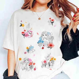 May include: Cream-colored t-shirt featuring colorful floral designs and illustrations of characters from a popular animated movie. The graphics include Elsa, Anna, Olaf, and other characters surrounded by flowers.