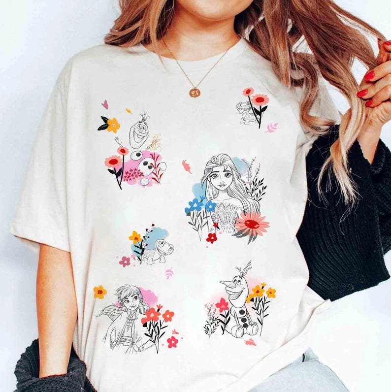 May include: Cream-colored t-shirt featuring colorful floral designs and illustrations of characters from a popular animated movie. The graphics include Elsa, Anna, Olaf, and other characters surrounded by flowers.