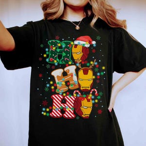 Puede incluir: Una camiseta negra de manga corta con un estampado gr&aacute;fico del texto HO HO HO. Las letras est&aacute;n decoradas con patrones verdes, texturas de pan de jengibre y rayas rojas y blancas. Tres cascos de Iron Man est&aacute;n integrados en el dise&ntilde;o.