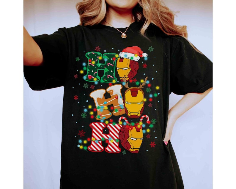 Puede incluir: Una camiseta negra de manga corta con un estampado gr&aacute;fico del texto HO HO HO. Las letras est&aacute;n decoradas con patrones verdes, texturas de pan de jengibre y rayas rojas y blancas. Tres cascos de Iron Man est&aacute;n integrados en el dise&ntilde;o.