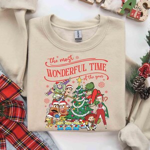 May include: Beige sweatshirt with a festive graphic featuring characters from a popular animated movie. The text "the most WONDERFUL TIME of the year" is printed in red. A Christmas tree and snowflakes are also included in the design.