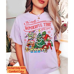 May include: A light purple t-shirt featuring a festive graphic with characters and a Christmas tree. The text reads "the most wonderful time of the year" in red. The shirt has a relaxed fit and short sleeves.