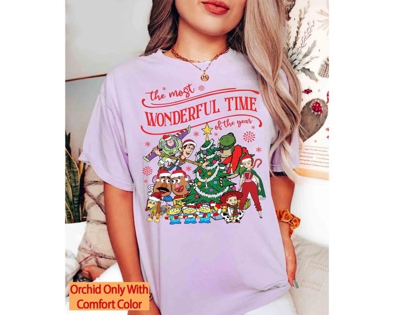 May include: A light purple t-shirt featuring a festive graphic with characters and a Christmas tree. The text reads "the most wonderful time of the year" in red. The shirt has a relaxed fit and short sleeves.