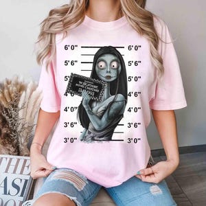 May include: Light pink t-shirt featuring a graphic of Sally from The Nightmare Before Christmas, holding a sign with text. The shirt has a crew neck and short sleeves. The background includes a height chart.