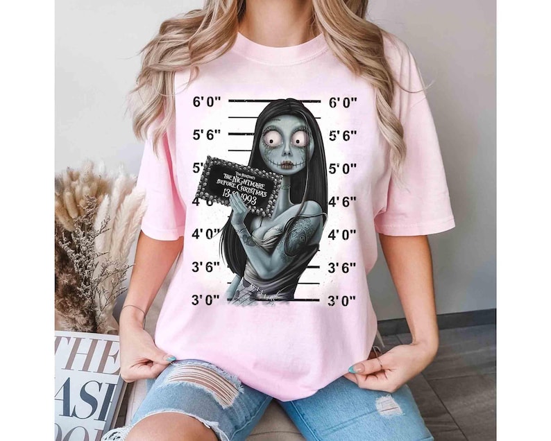 May include: Light pink t-shirt featuring a graphic of Sally from The Nightmare Before Christmas, holding a sign with text. The shirt has a crew neck and short sleeves. The background includes a height chart.