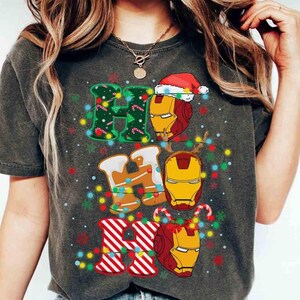 Puede incluir: Una camiseta gris carb&oacute;n con el texto HO HO HO. La primera H es verde con bastones de caramelo, la O es un casco de Iron Man con un gorro de Pap&aacute; Noel, la segunda H es de pan de jengibre, la segunda O es un casco de Iron Man, la tercera H es de rayas rojas y blancas, y la tercera O es un casco de Iron Man con astas de reno, todo rodeado de luces navide&ntilde;as de colores.