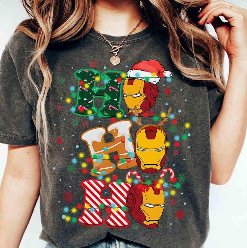 Puede incluir: Una camiseta gris carb&oacute;n con el texto HO HO HO. La primera H es verde con bastones de caramelo, la O es un casco de Iron Man con un gorro de Pap&aacute; Noel, la segunda H es de pan de jengibre, la segunda O es un casco de Iron Man, la tercera H es de rayas rojas y blancas, y la tercera O es un casco de Iron Man con astas de reno, todo rodeado de luces navide&ntilde;as de colores.
