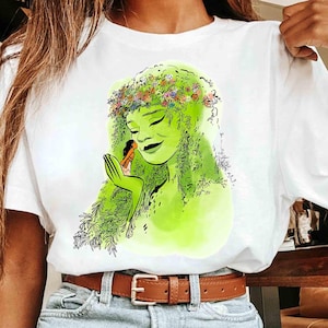 May include: White t-shirt featuring a green illustration of a woman with a floral crown and flowing green hair. The woman is holding a small figure in her hand. The design has a watercolor effect.