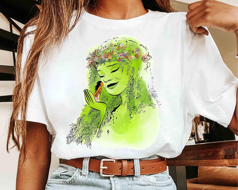 May include: White t-shirt featuring a green illustration of a woman with a floral crown and flowing green hair. The woman is holding a small figure in her hand. The design has a watercolor effect.