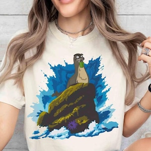 May include: Cream-colored t-shirt featuring a cartoon seal holding a green cup, perched on a rock amidst blue and white waves. The seal has a surprised expression. The design is printed on a soft, light-colored tee.