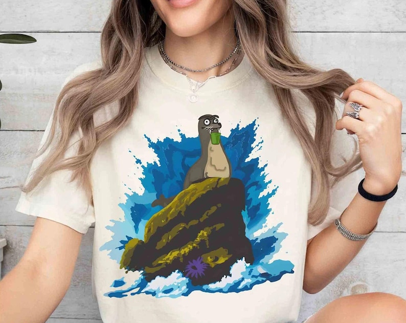 May include: Cream-colored t-shirt featuring a cartoon seal holding a green cup, perched on a rock amidst blue and white waves. The seal has a surprised expression. The design is printed on a soft, light-colored tee.