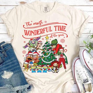 May include: Cream-colored t-shirt featuring a festive design with the text "The Most Wonderful Time of the Year" in red. The graphic includes cartoon characters, a Christmas tree, and snowflakes, creating a holiday theme.